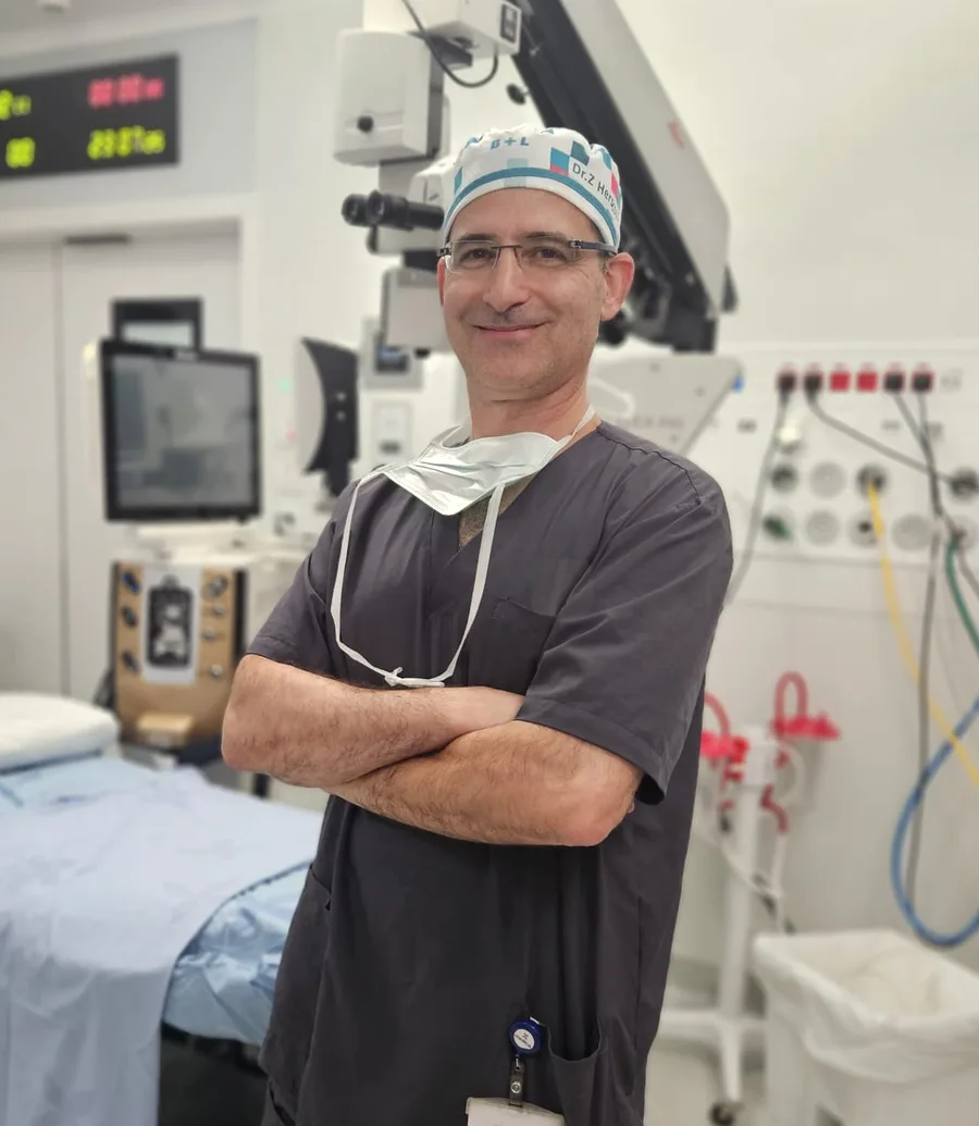ד״ר צבי הרשקוביץ בחדר הניתוח, ידיים שלובות, לצד מיקרוסקופ הניתוח