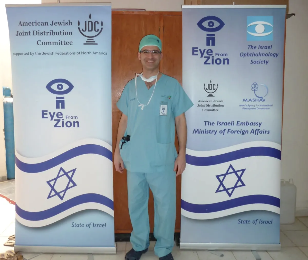 ד״ר הרשקוביץ בבגדי ניתוח בין שני שלטי משלחת Eye From Zion עם דגלי ישראל